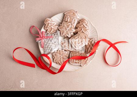 Festive assortment of homemade Christmas gingerbread cookies shaped as houses, trees and snowflakes on a white plate, decorated with a bright red ribb Stock Photo