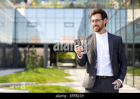 Businessman smiling while checking content on his mobile phone, standing outside a contemporary glass office building, representing modern communication and business success Stock Photo