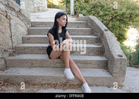 A young woman sits on concrete stairs outdoors, looking to the side in a quiet moment. Stock Photo