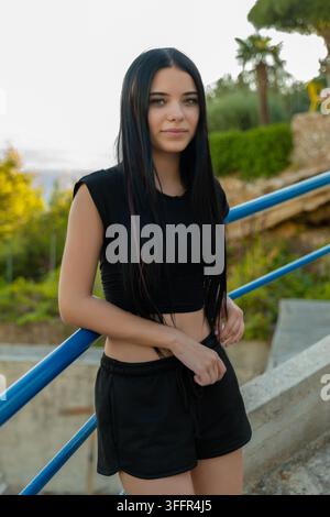 Young woman in a black outfit stands outdoors near blue railings with a calm expression. Stock Photo