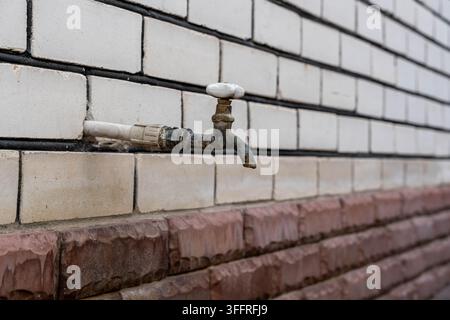 Rusty outdoor faucet on tiled wall shows weathered plumbing in close up. Old metal tap has white handle, corroded pipe, and brick masonry backdrop  Stock Photo