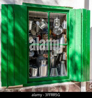 Green wooden window with its shutters open, revealing an assortment of metal jugs, pitchers, and kitchenware neatly arranged inside. Stock Photo
