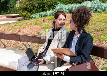 Businesswomen collaborating on park bench discussing work outdoors Stock Photo