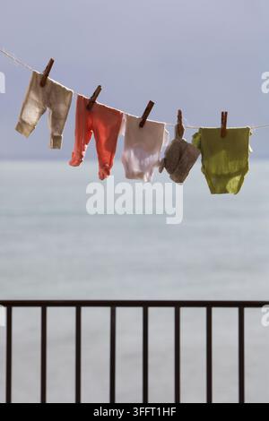 Baby clothing air drying on an outdoor clothesline, above a dark metal balcony railing with a scenic ocean and sky view. Stock Photo