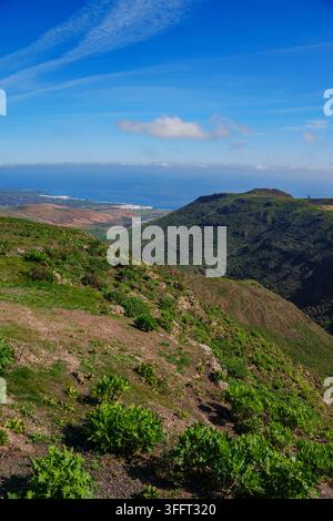 Rolling hills covered in green shrubs stretch toward distant blue ocean. Sky above is bright blue with wispy clouds. Lanzarote Road cycling Stock Photo