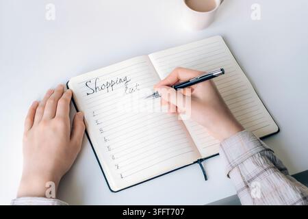 Person writing a shopping list in an open notebook with a pen, organized planning and everyday household budgeting on a clean white desk. Top view. Stock Photo