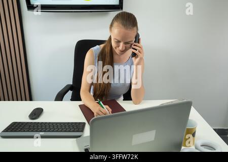 Young woman multitasking with phone call, writing notes, and working on laptop at her desk Stock Photo