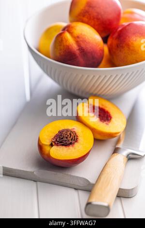 Bright, ripe nectarines arranged in a white bowl with a halved, juicy peach on a cutting board beside a wooden-handled knife, evoking fresh summer fru Stock Photo