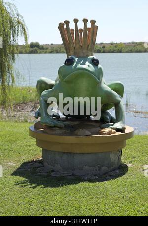 Green frog king sculpture wearing a golden crown on a pedestal by a lakeside in a sunny park, surrounded by grass and willow trees. Bulgaria, Balchik, Stock Photo