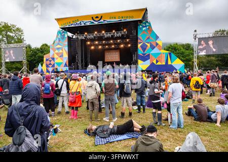 FESTIVAL CROWD RELAXING ON THE GRASS, REPORTAGE, GREEN MAN FESTIVAL 2015, BRECON, WALES, UK, 2015: A person wearing a black hoodie and shorts lies resting on a blue plaid blanket on the grass, while other members of the audience stand and watch the main stage. Photo: Rob Watkins. INFO: The Green Man Festival is held annually in the Brecon Beacons in August. It celebrates alternative music, arts, literature, science and family-friendly culture in a beautiful mountain setting. The 2015 event started sunny but turned muddy with overnight rain on the final two days. Stock Photo