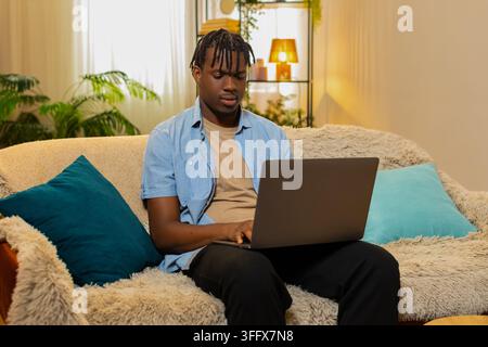 African American young man on home sofa works on laptop checking text and fixing small details with focused attention. Black guy on home sofa edits a document and reviews work with calm concentration. Stock Photo