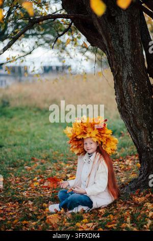 A charming ginger girl in a large autumn leaf crown sits cross-legged on the ground under a big tree, smiling and looking into the camera on a beautif Stock Photo