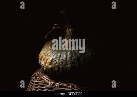 Single green pumpkin on wicker basket in dark low key lighting with deep shadows. Minimal autumn still life with rustic texture and harvest mood Stock Photo