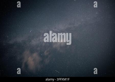 Starry night sky with Milky Way galaxy and faint meteor streaks behind light clouds Stock Photo