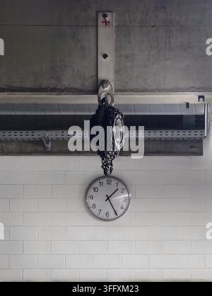 A round analog clock with black hands and numbers suspended by a heavy metal chain from a ceiling mount against a white brick wall in a minimalist ind Stock Photo