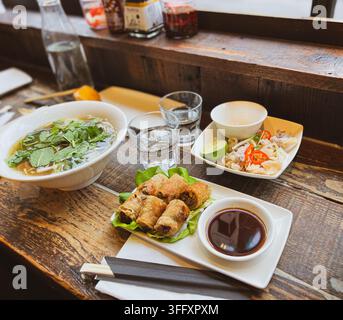 A wooden table spread with Vietnamese dishes including pho noodle soup, spring rolls and salt and pepper squid, England, UK Stock Photo