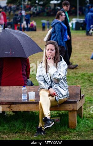 WOMAN IN SILVER JACKET SMOKING AT A RAINY FESTIVAL, REPORTAGE, GREEN MAN FESTIVAL 2015, BRECON, WALES, UK, 2015: A young woman wearing a shiny silver jacket and metallic gold leggings smokes a cigarette while sitting on a wet wooden bench at a rainy outdoor festival. Photo: Rob Watkins. INFO: The Green Man Festival is held annually in the Brecon Beacons in August. It celebrates alternative music, arts, literature, science and family-friendly culture in a beautiful mountain setting. The 2015 event started sunny but turned muddy with overnight rain on the final two days. Stock Photo