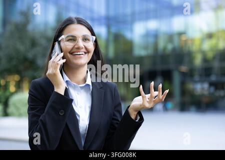 Young businesswoman smiling while communicating on her mobile phone, having a successful conversation during a break outside a modern corporate building Stock Photo