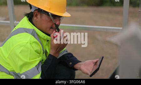 Close-up shot of an Asian male engineer wearing safety hardhat and vest using a walkie talkie radio and checking a smartphone at an outdoor industrial Stock Photo