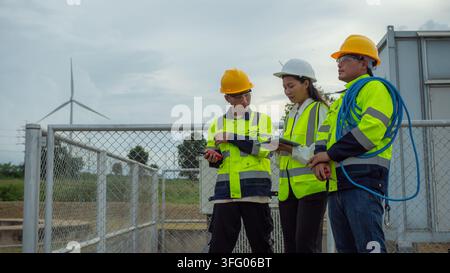Three Asian electrical engineers, two men and one woman in hardhats and safety vests, inspect wind turbine power plant outdoor using digital tablet. Stock Photo