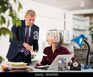 Man in suit shouting at senior woman who sitting at table in office because of documentation truble. Stock Photo