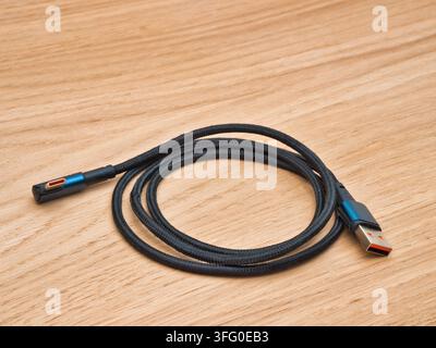 Black Braided USB Cable on Wood Surface - A black braided USB-C to USB-A data and charging cable is coiled on a natural light wooden background, ideal Stock Photo