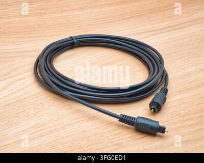 Black Optical Audio Cable on Wood - A black TOSLINK optical audio cable with gold-tipped connector coiled neatly on a light wooden background, ready f Stock Photo