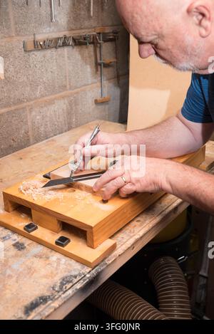 Man marking wood on a workbench. Wood shavings are visible. He wears a blue shirt and a ring. Tools hang on a concrete wall. Stock Photo