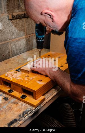 Man drilling wood on a wooden workbench. He wears a blue shirt and a ring. Wood shavings are visible. Concrete block wall in background. Stock Photo