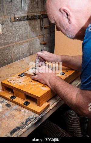 A bald man in a blue shirt marks a small wooden piece with a pencil on a workbench. Wood shavings are present. A concrete wall and tools are in the ba Stock Photo