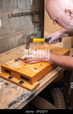 A bald man hammers a nail into a piece of wood on a workbench. Wood shavings are scattered. Tools hang on the concrete wall. A dust collection hose is Stock Photo
