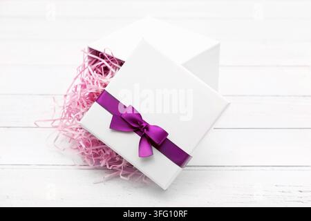 Elegant white gift box opened with gold ribbon bow and beige shredded paper filler on a white wooden background, perfect for luxury packaging. Stock Photo