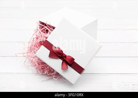 Elegant white gift box opened with gold ribbon bow and beige shredded paper filler on a white wooden background, perfect for luxury packaging. Stock Photo