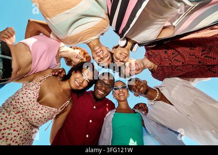 Diverse group of happy young people standing in a circle, looking down at camera, smiling, against clear blue sky Stock Photo