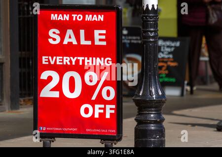 A vibrant red A-board outside a clothing store promotes a "MAN TO MAN SALE: 20% OFF" on the busy high street beside a black metal post. Southport, UK. Stock Photo