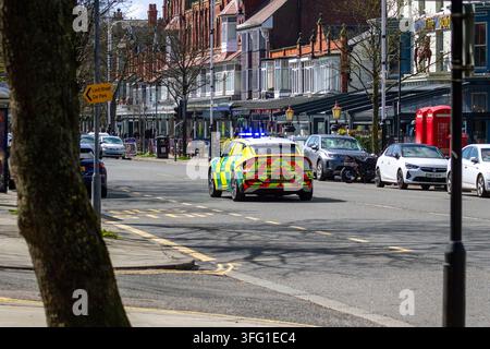 A North West Ambulance Service paramedic vehicle with flashing blue lights speeds down a historic street full of shops and parked cars. Southport, UK. Stock Photo