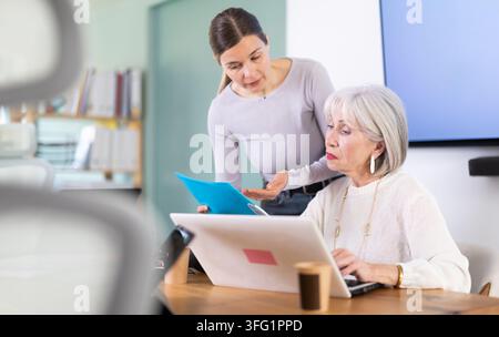 Elderly woman works on laptop while adult colleague shows folder with documents in office Stock Photo