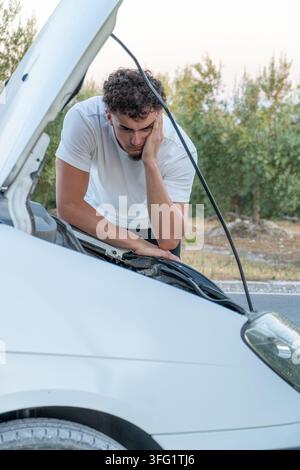 Young man leaning on car engine, experiencing stress and frustration after vehicle breakdown on the side of a road Stock Photo