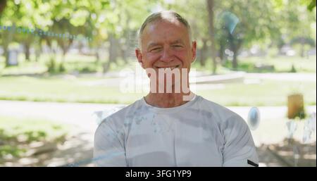 Posing senior man wearing light longsleeve shirt smiling, folding arms in sunny park with trash bin Stock Photo