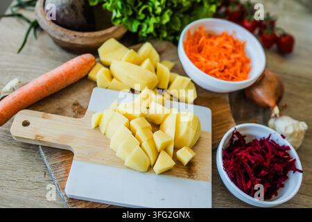 Fresh vegetables are arranged on a kitchen counter. Potatoes, carrots, and beets are cut and placed on a wooden board. Other veggies are in bowls near Stock Photo