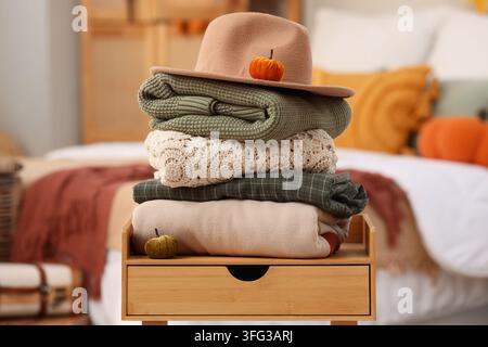 Autumn comfort and seasonal transition, stack of warm sweaters and felt hat with decorative pumpkins on a wooden bedside table in a cozy bedroom Stock Photo
