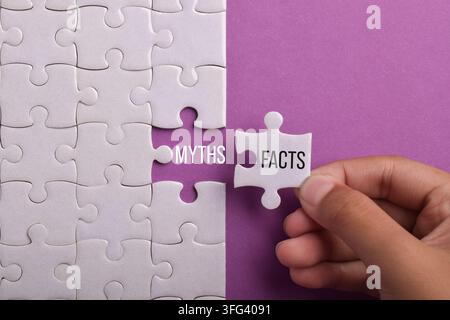 Hand placing a facts jigsaw piece beside myths on a white puzzle over a purple background, representing truth, misinformation, critical thinking Stock Photo