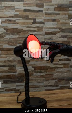 Black robotic or Prosthesis hand reaches toward a glowing yellow bulb in a modern desk lamp against a stone wall, useful for concepts of AI, innovatio Stock Photo