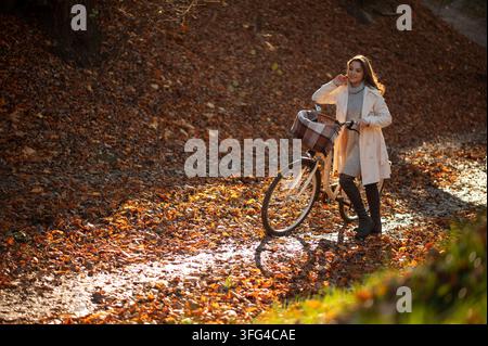 A smiling young woman stands with her bicycle on a path covered in fallen leaves, bathed in warm sunlight Stock Photo