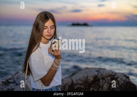 Young Woman Using Smartphone by Sea at Sunset. Young woman looks at a smartphone while standing beside a rocky seashore at sunset with calm water and Stock Photo
