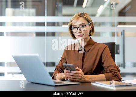 Professional businesswoman with eyeglasses at office desk holding smartphone, looking at camera modern digital work, corporate communication, connectivity and focused leadership Stock Photo