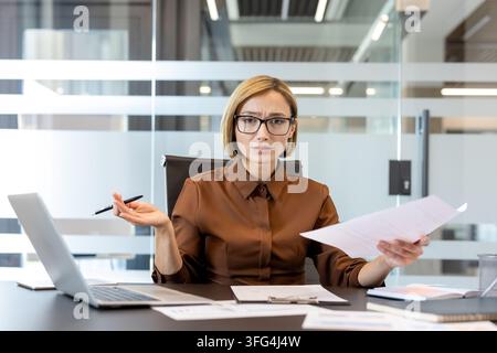 Businesswoman sitting at office desk, holding documents, and gesturing with an open hand, expressing confusion and frustration while dealing with overwhelming paperwork and digital tasks Stock Photo