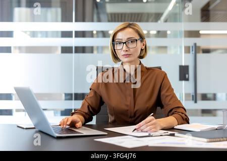 Professional businesswoman wearing glasses typing on laptop and writing on documents, performing various tasks related to her career in a modern office environment Stock Photo