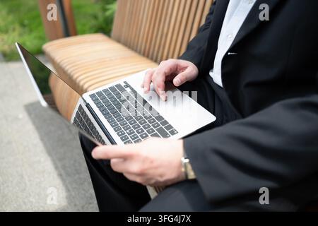 Close up of businessman using laptop while sitting on outdoor bench. Mobile office, remote work, business technology and flexible working concept. Stock Photo