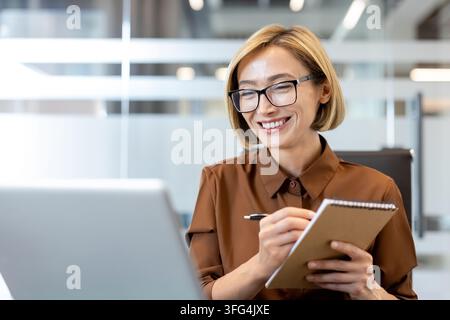 Smiling businesswoman in glasses sitting at her desk, working on a laptop while taking notes in a spiral notepad, showing focus, organization, and a positive professional attitude Stock Photo